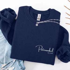 Puede incluir: Una sudadera azul marino de cuello redondo con texto bordado en blanco que dice Patenonkel Est. 2025 en la parte inferior derecha. La prenda tiene mangas largas con pu&ntilde;os acanalados y una etiqueta de la marca Gildan en el cuello.