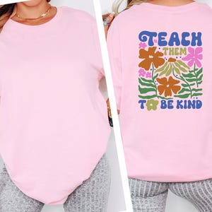 May include: A light pink t-shirt with the words "Teach Them To Be Kind" in a colorful floral design on the back. The shirt is a solid color and has a crew neck.