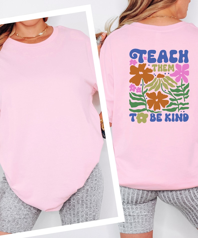 May include: A light pink t-shirt with the words "Teach Them To Be Kind" in a colorful floral design on the back. The shirt is a solid color and has a crew neck.