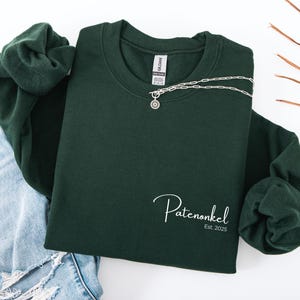Puede incluir: Una sudadera de cuello redondo en color verde bosque oscuro con el texto en cursiva blanca Patenonkel y el texto impreso Est. 2025 en la parte inferior derecha. Un collar de cadena plateada con un colgante circular descansa en el cuello.