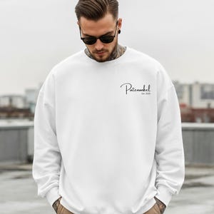 Puede incluir: Una sudadera blanca de manga larga con cuello redondo que presenta el texto negro Patenonkel Est. 2025 impreso en una fuente cursiva en la parte superior izquierda del pecho. La prenda tiene un corte est&aacute;ndar con pu&ntilde;os y cuello acanalados.