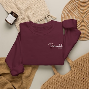 Puede incluir: Una sudadera de cuello redondo y manga larga en color burdeos intenso, con un texto bordado en blanco en el pecho izquierdo que dice Patenonkel Est. 2025 en una fuente cursiva. La prenda tiene un cuello redondo sencillo y pu&ntilde;os acanalados.