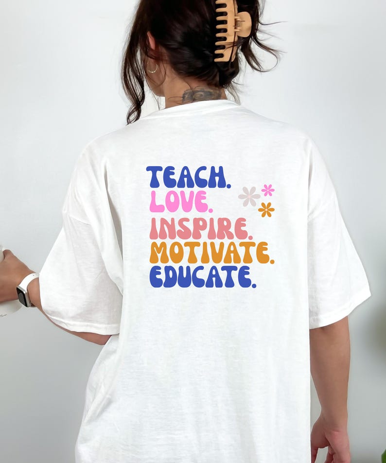 May include: White t-shirt with the words "TEACH. LOVE. INSPIRE. MOTIVATE. EDUCATE." in blue, pink, orange, and blue lettering. Two small flower graphics are to the right of the text. The shirt is a casual style.
