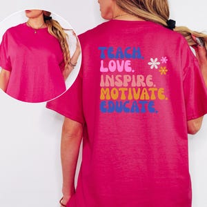 May include: A vibrant fuchsia t-shirt with short sleeves. The back of the shirt features the words "TEACH, LOVE, INSPIRE, MOTIVATE, EDUCATE" in a colorful, retro font, with two small flower graphics. A casual, comfortable tee.