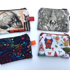 May include: Four small zippered pouches with various patterns. One features a skull illustration, another a floral design with a skull, a third has colorful cat-themed text, and the last showcases garden gnomes and flowers. Each pouch has a colored tab.
