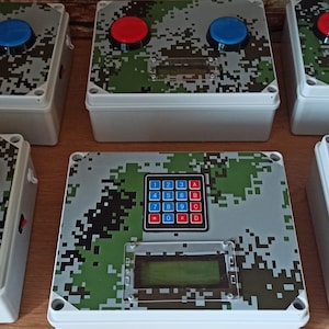Pode incluir: Várias caixas brancas com tampas estampadas com camuflagem. Cada caixa tem um botão vermelho e azul, um pequeno ecrã, e algumas têm um teclado. Estão dispostas sobre uma superfície de madeira.