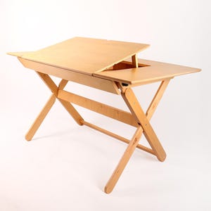 May include: A light-colored wooden drafting table with an adjustable top. The table has a rectangular surface with a section that can be raised. The legs are in an X-shape, providing stability.