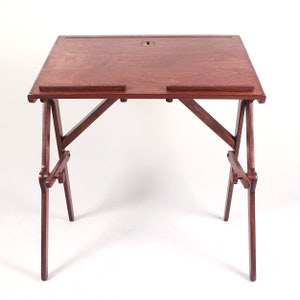 May include: A dark wood folding desk with a simple design. The desk has a rectangular top with a small hole in the center and two small shelves on the front. The legs are made of wood and fold up for easy storage.