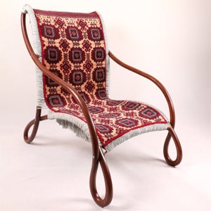 May include: A wooden armchair with a red and cream patterned fabric seat and back. The fabric has a fringe border. The chair has a unique curved design.