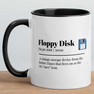 May include: White ceramic mug with a black interior and handle. The mug features the words "Floppy Disk" with a definition and a graphic of a floppy disk. The text describes it as a vintage storage device.