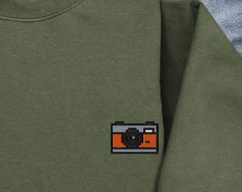 May include: An olive green sweatshirt with a pixelated camera design on the chest. The camera is outlined in black with orange and gray details. The sweatshirt is made of a soft material and has a crew neck.