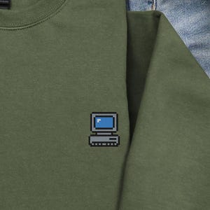 Puede incluir: Sudadera verde oliva con un gráfico de ordenador pixelado en el pecho. La sudadera de cuello redondo está hecha de un material suave y es de estilo casual. El diseño es un ordenador retro.