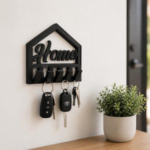 May include: A black wall-mounted key holder shaped like a house outline with the word Home in cursive script. The holder features five downward-facing hooks, with three sets of metal key rings and car keys hanging from the hooks on the wall.