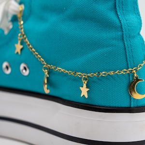 May include: A pair of turquoise blue canvas shoes with a gold chain ankle bracelet featuring star and crescent moon charms.