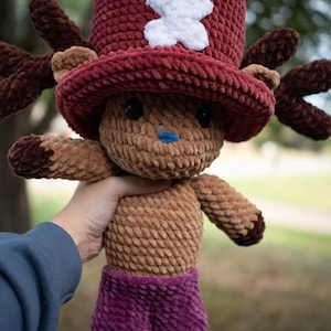 May include: A plush crochet doll of a character with brown fur, a blue nose, and dark eyes. The doll wears a large, textured burgundy top hat featuring a white cross emblem and brown antlers on the sides. It is dressed in purple shorts.