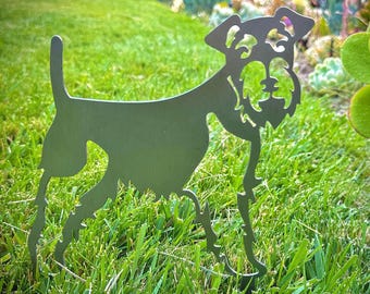 May include: A metal silhouette of a dog, possibly a miniature schnauzer, standing in a grassy area. The silhouette is a dark green colour and is detailed with the dog's facial features.