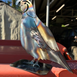 May include: A metal sculpture of a quail with an iridescent finish. The sculpture is standing on a black metal base.