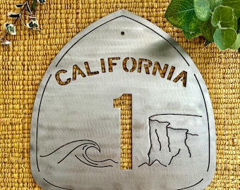 May include: A metal sign featuring a wave and a cliff design. The sign is shaped like a California highway sign and reads "CALIFORNIA 1" in a cut-out font.