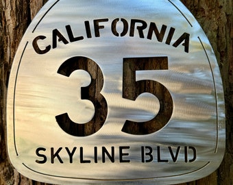 May include: A metal sign with a California route shield design. The sign is silver with black lettering and features the text "CALIFORNIA 35 SKYLINE BLVD".