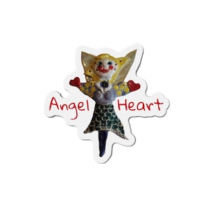 May include: A whimsical sticker featuring a ceramic angel with blonde hair, red heart-shaped hands, and a green scale-patterned skirt. The text "Angel Heart" is written in red script across the sticker.