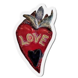 May include: A red heart-shaped ceramic ornament with the word "LOVE" written in gold lettering. The heart has a smaller black heart in the center and a textured, blue and green design at the top.