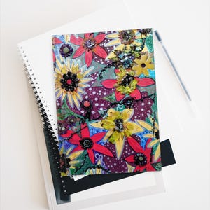 May include: A spiral-bound notebook with a colorful floral design. The cover features red, yellow, and purple flowers with black accents on a patterned background. The notebook is stacked on top of other notebooks and a pen.
