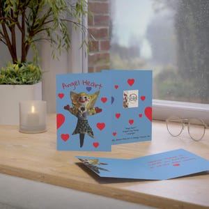 May include: A light blue greeting card with the words "Angel Heart" and a whimsical angel figure. The card features red hearts and a small image of the artist's signature. The card is open, revealing a message inside.