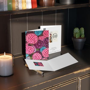May include: A greeting card with a colorful floral design featuring pink, purple, and blue circles. The card is open, revealing a blank interior and a small square image of the design. A lit candle and a pen are in the foreground.