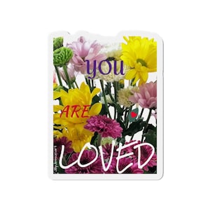 May include: A sticker featuring a vibrant bouquet of yellow, pink, and purple flowers. The words "YOU ARE LOVED" are written across the flowers in a handwritten font. The sticker has a white border.