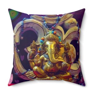 May include: A gold-colored statue of Ganesha, the Hindu god of wisdom and prosperity, sits in front of a carved wooden Om symbol. The statue is adorned with colorful beads and a garland of flowers. The background is a purple and pink ombre with hearts and a mandala.