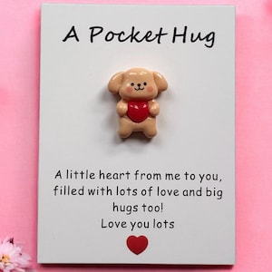 Puede incluir: Tarjeta "A Pocket Hug" con una pequeña figura de perro beige sosteniendo un corazón rojo. La tarjeta incluye el texto: "Un pequeño corazón de mí para ti, lleno de mucho amor y grandes abrazos también! Te quiero mucho."