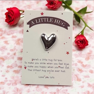 以下が含まれることがあります： 「A LITTLE HUG」の文字が入ったカードに、銀色のハート型のチャーム。カードは星で装飾され、「Here's a little hug for you. Love you lots.」の文字。背景には赤いバラが配置されています。