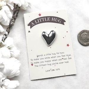 May include: A silver heart-shaped pin on a white card with the text "A LITTLE HUG". The card includes a message about hugs and is decorated with red stars and dots. A coin is visible in the background.