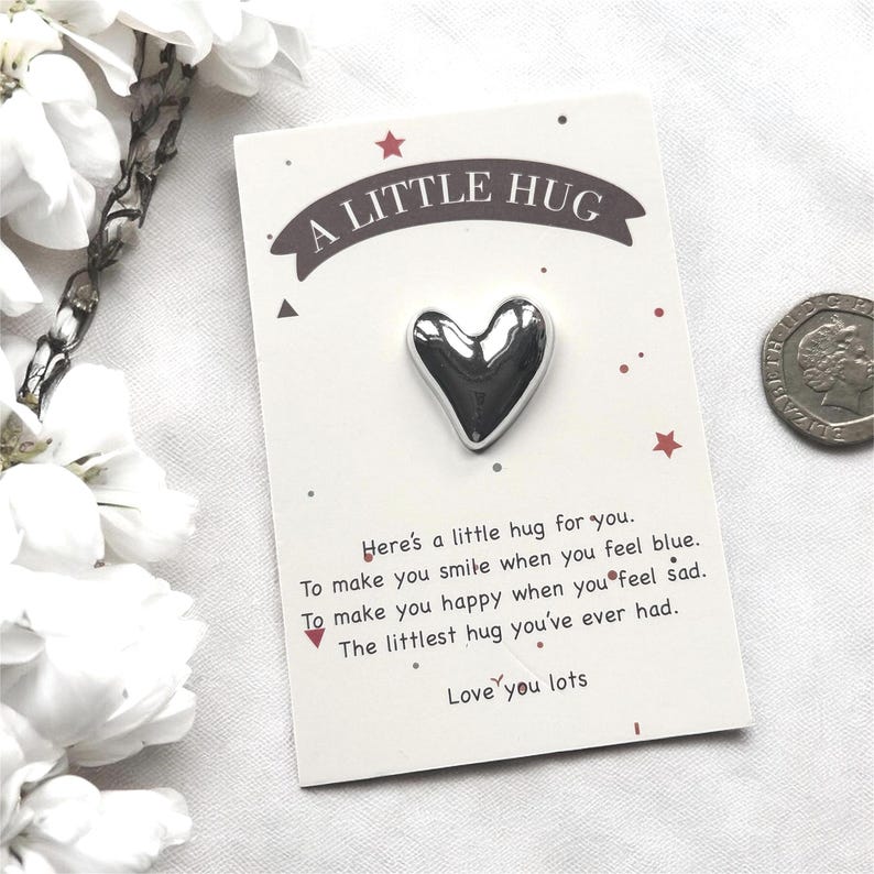 以下が含まれることがあります： 「A LITTLE HUG」の文字が入った白いカードに、銀色のハート型のピン。カードにはハグに関するメッセージが書かれており、赤い星と点で装飾されています。背景にはコインが見えます。