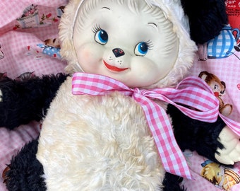 May include: A vintage panda bear plush toy with a white face, black ears, and a cream-colored body. The panda has blue eyes, a red mouth, and a pink gingham ribbon. The plush is set against a pink gingham background.