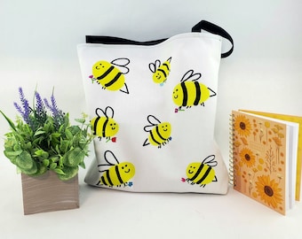 May include: White tote bag with a black handle and yellow and black bee illustrations. The bees are drawn in a simple, cartoon style and have smiling faces. The bag is perfect for carrying groceries, books, or other items.
