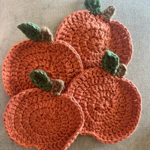 May include: Four handmade crochet pumpkin coasters in varying sizes. The coasters are orange with green and brown accents, resembling miniature pumpkins. The coasters are arranged on a neutral-colored surface.
