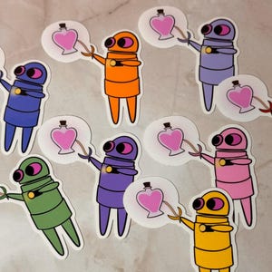 May include: Nine colorful robot stickers with pink hearts and bottles. The robots are in different colors, including red, orange, yellow, green, blue, purple, and pink.