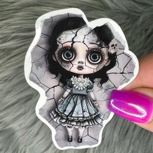 May include: A sticker featuring a cartoon illustration of a cracked porcelain doll with large eyes, wearing a blue dress with white lace trim. The doll is set against a cracked background.