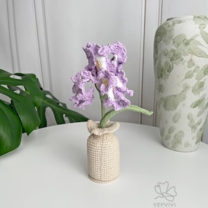 May include: A handcrafted crochet flower arrangement featuring a lavender iris with white and yellow accents, displayed in a beige vase. The flower has green leaves and is placed on a white surface.
