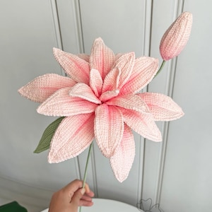 May include: A large, handmade crochet flower in shades of pink, with a green stem and leaf. The flower has multiple petals and a bud. The craftwork is detailed, and the flower is held by a hand. The flower is a decorative item.