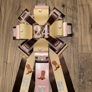 May include: A decorative rosette with brown, tan, and pink ribbons. The number "24" is in the center, with the letters "H A P P Y B D A Y" on the ribbons. Cowboy boot embellishments are also present.