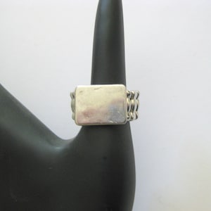 May include: A silver-toned ring with a rectangular face and a textured band. The ring is displayed on a black mannequin hand against a white background. The ring's design features a series of parallel lines on the sides.