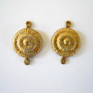 May include: Two gold-toned metal connector pieces with a circular design. Each piece has a raised, textured centre with a smaller circle in the middle. The pieces have two small loops on the top and bottom.