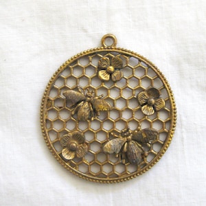 May include: A gold-toned metal pendant featuring a honeycomb design with three bees and four flowers. The pendant is round and has a loop at the top for hanging.