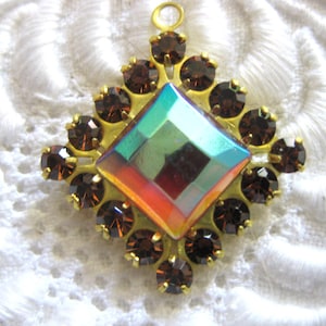 May include: A vintage pendant with a central iridescent square stone, surrounded by brown rhinestones and a gold-tone metal setting. The pendant has a small loop at the top for hanging.