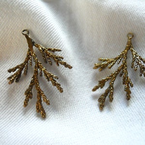 May include: Two gold-toned metal charms shaped like a branch of a coniferous tree. The charms have a loop at the top for attaching to jewelry or other crafts.
