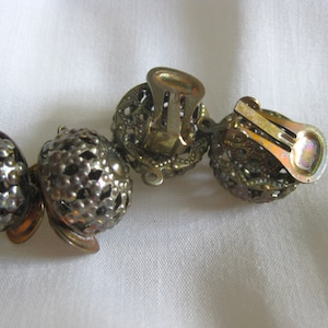 May include: A pair of gold-toned clip-on earrings with a round, openwork design. The earrings have a textured surface and are decorated with small, raised dots.
