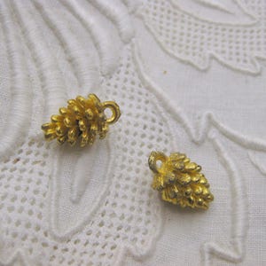 May include: Two small, gold-colored pinecone charms with textured surfaces and loop attachments. They rest on a white, patterned fabric background. These charms are suitable for jewelry making or craft projects.