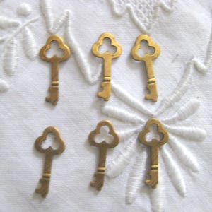 May include: Six small, antique-style keys with a decorative trefoil design. The keys are made of a gold-colored metal and are arranged on a white, embroidered fabric. The keys have a vintage aesthetic.